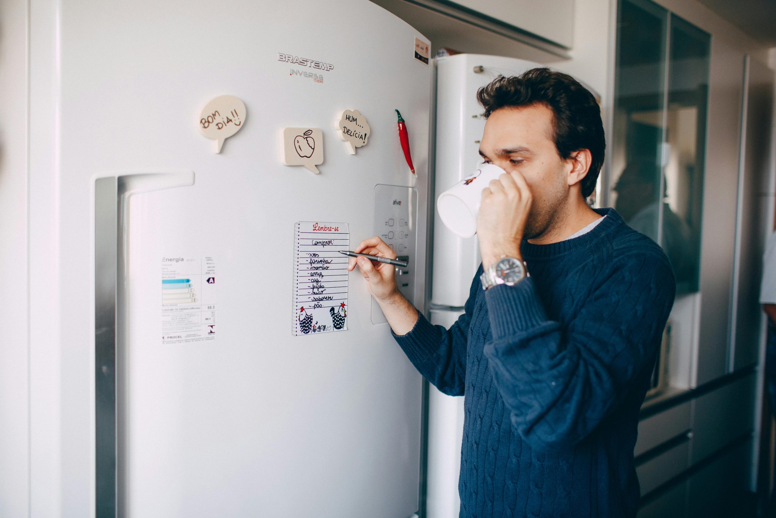 Home Man drinks coffee while writing a to-do list on a kitchen refrigerator.